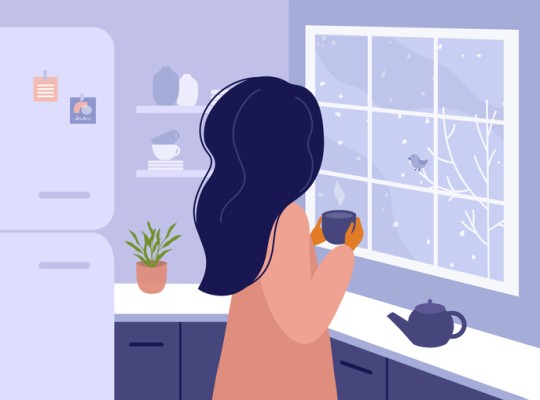 Woman with a cup of tea or coffee in her hands standing in the kitchen at home and looking out the window