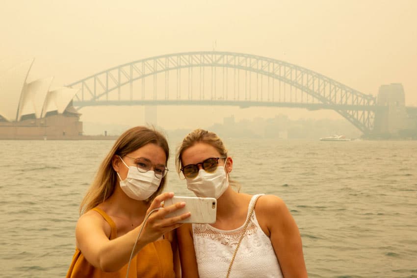 Two white women wearing surgical masks and taking a selfie in front of Sydney Harbor Bridge obscured by bushfire smoke